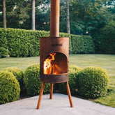 Rust-colored metal chiminea with flames on a patio surrounded by greenery