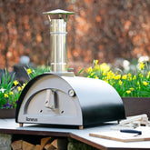Outdoor pizza oven with 'Igneus' branding on a wooden table with flowers and logs in the background.