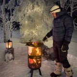 Person using a portable fire pit in a snowy forest setting with string lights on trees.