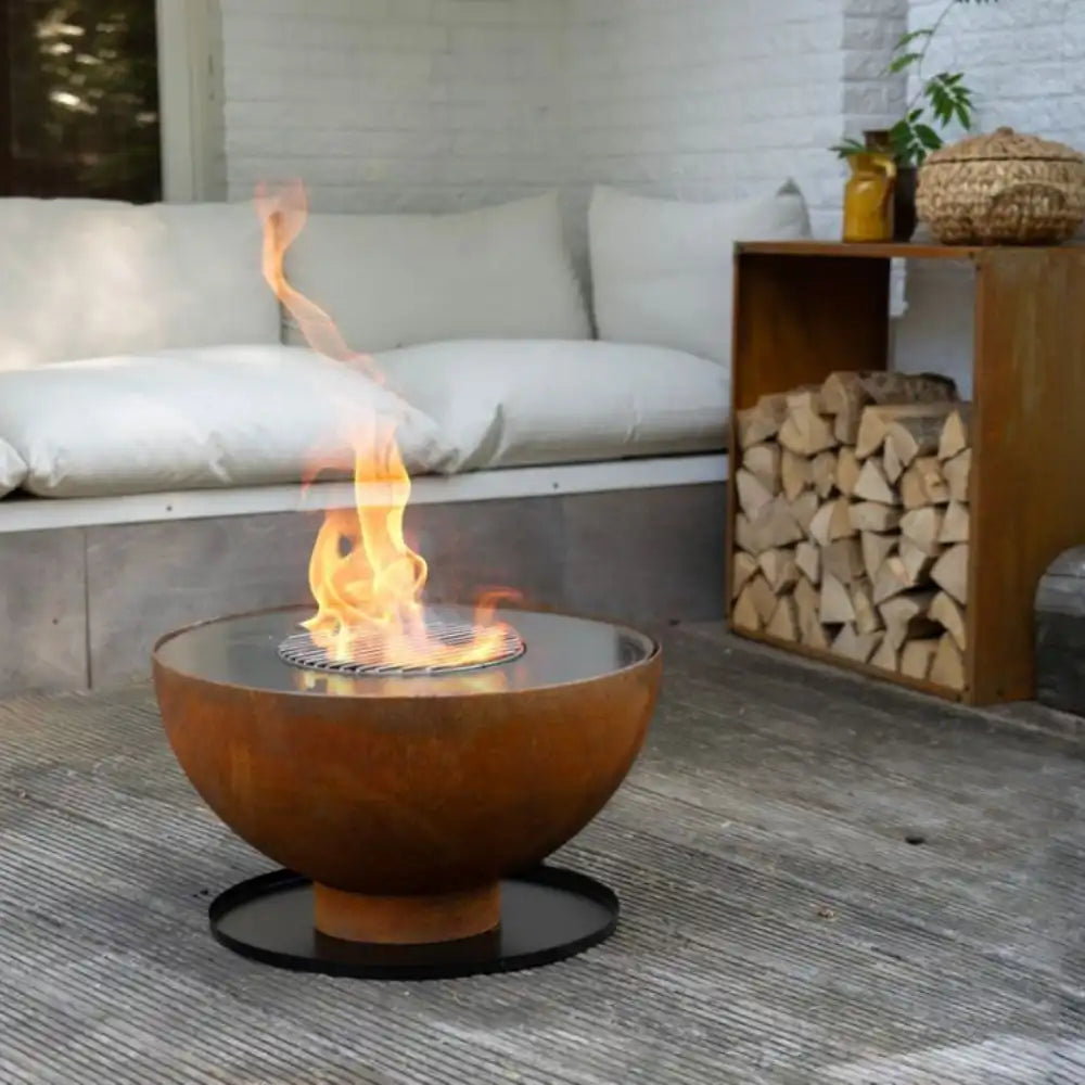 Outdoor fire pit table with flames on a patio with a couch and stacked logs in the background.
