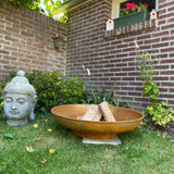 Rust-colored fire pit with wooden logs in a garden setting with a Buddha statue and brick wall.