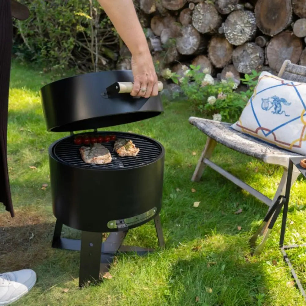 Person grilling on a small black barbecue grill outdoors with a bench and logs in the background.
