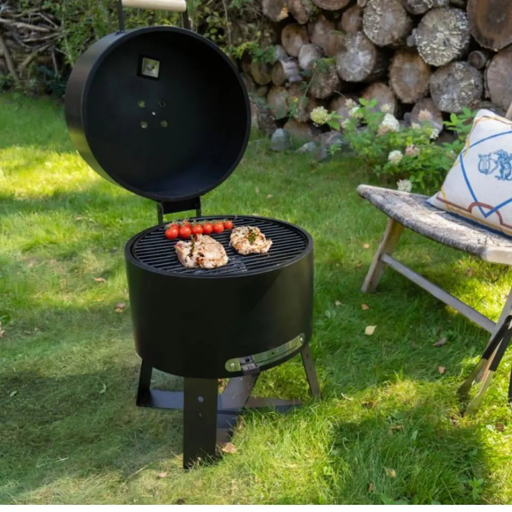 Black portable grill with food on a grassy area with logs and a chair in the background