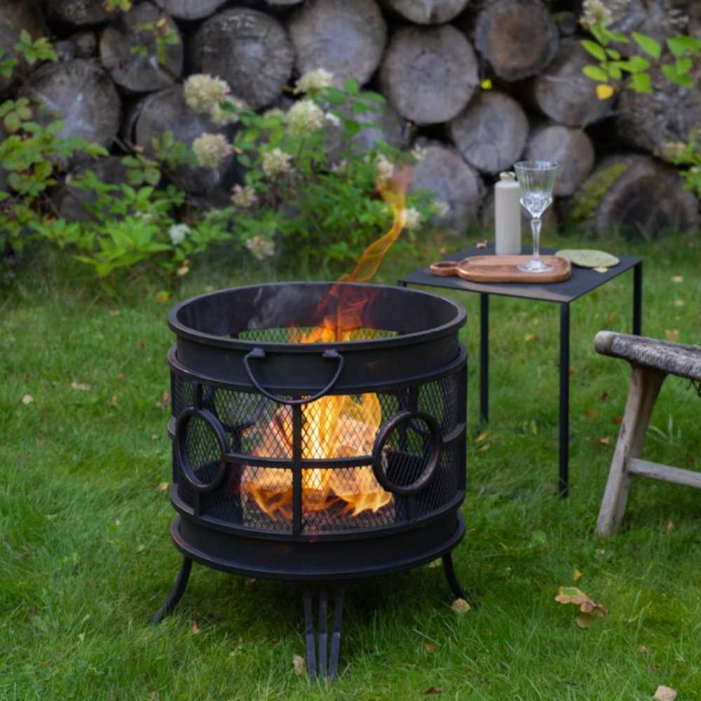 Black metal fire pit with flames in a garden setting, surrounded by logs and a small table with items.