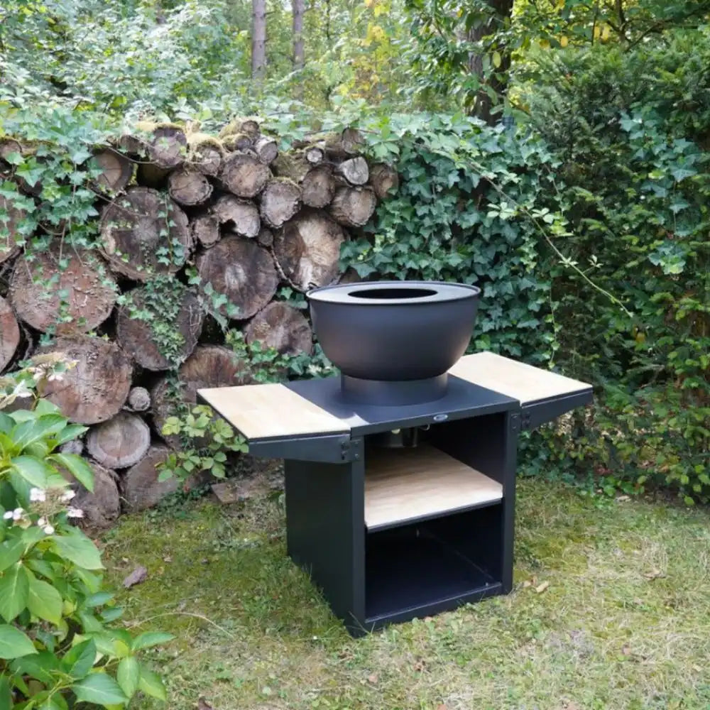 Black fire pit table with a bowl on top in a garden setting with logs and greenery.