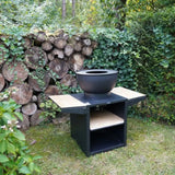 Black fire pit table with a bowl on top in a garden setting with logs and greenery.