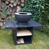 Black outdoor fire pit with a bowl on top, surrounded by stacked logs and greenery.