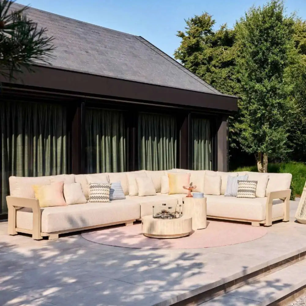 Outdoor patio with beige sectional sofa, cushions, and a small fire pit in a garden setting.