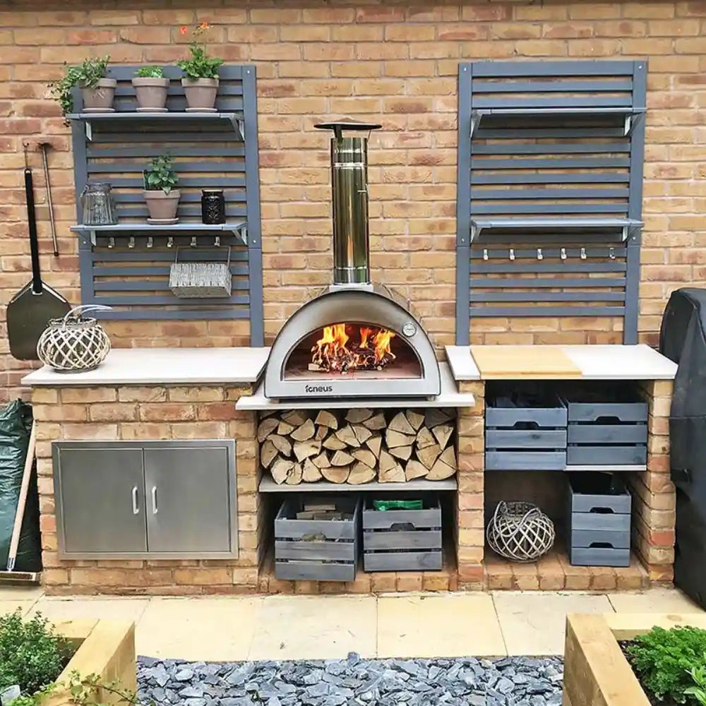 Outdoor kitchen setup with pizza oven, shelves, and storage units against a brick wall.