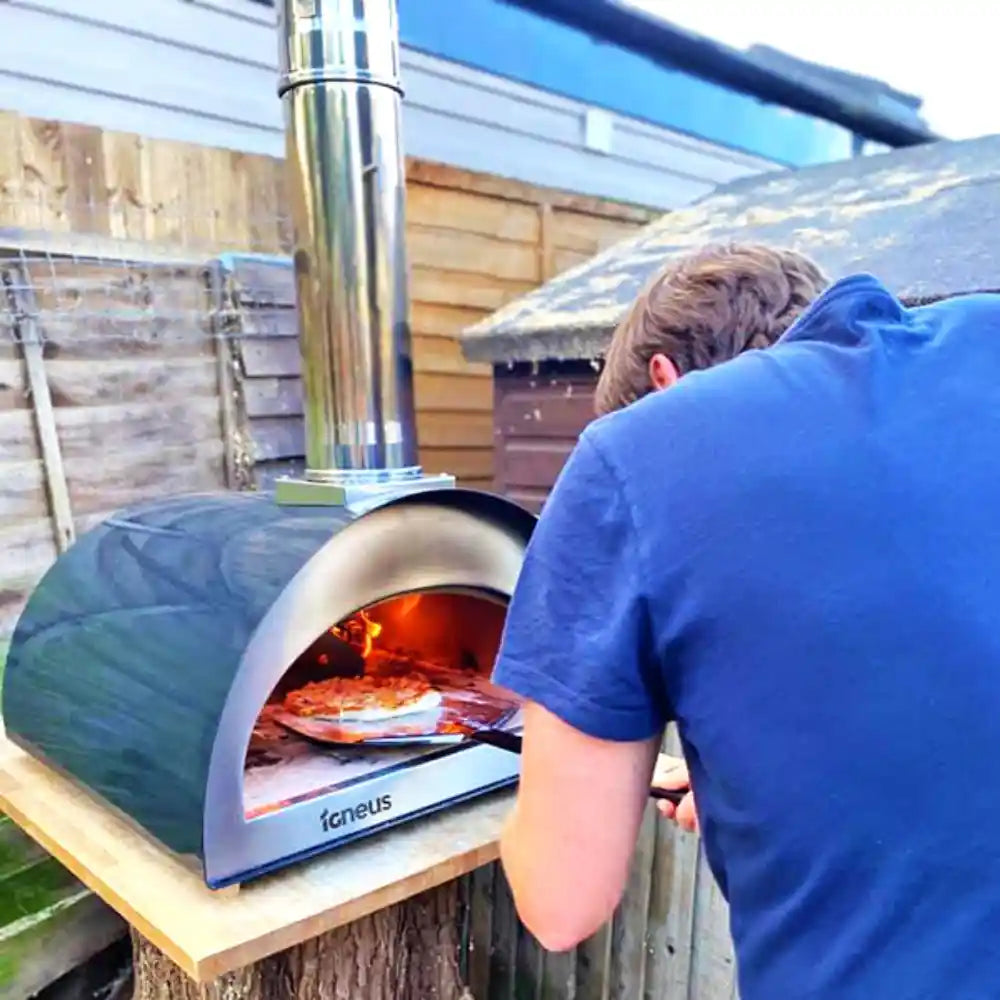 Person cooking pizza in an outdoor pizza oven branded 'Igenius' on a wooden table.