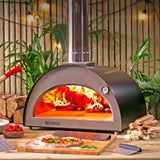 Outdoor pizza oven with flames, surrounded by plants and wood, on a wooden surface.