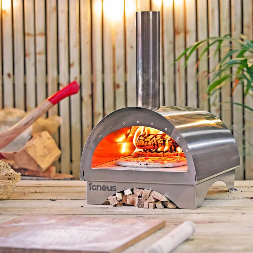 Outdoor pizza oven with wood fire cooking a pizza, surrounded by wooden logs and plants.