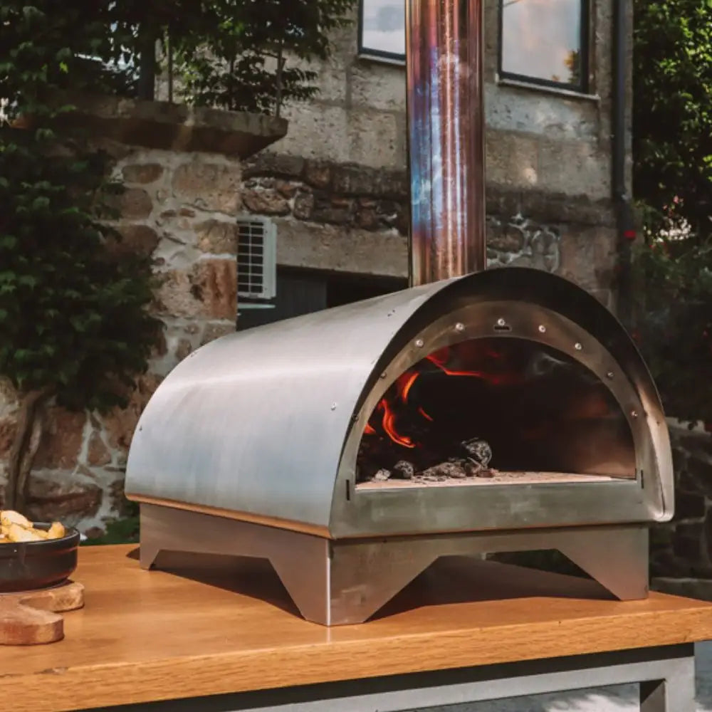 Metal outdoor pizza oven with fire inside, placed on a wooden surface outdoors.