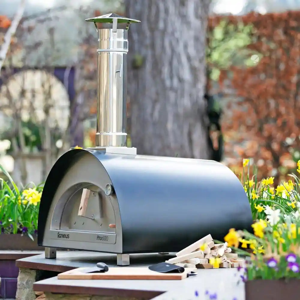 Outdoor pizza oven with wood and accessories on a patio with flowers and trees in the background