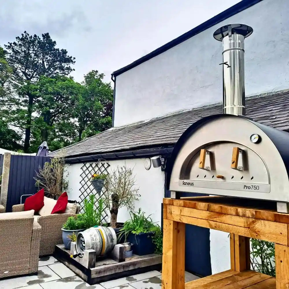 Outdoor pizza oven on a wooden stand with a white building and garden in the background