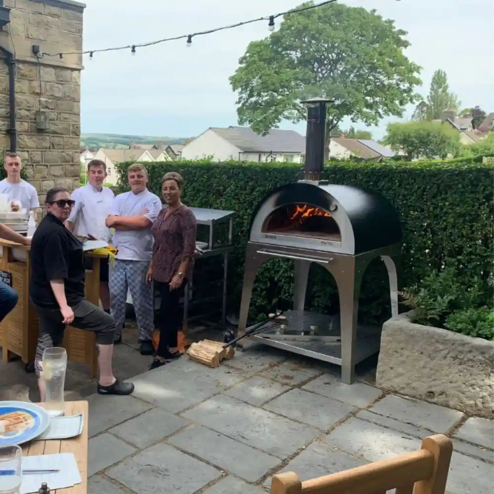 Group of people gathered around an outdoor pizza oven in a garden setting.