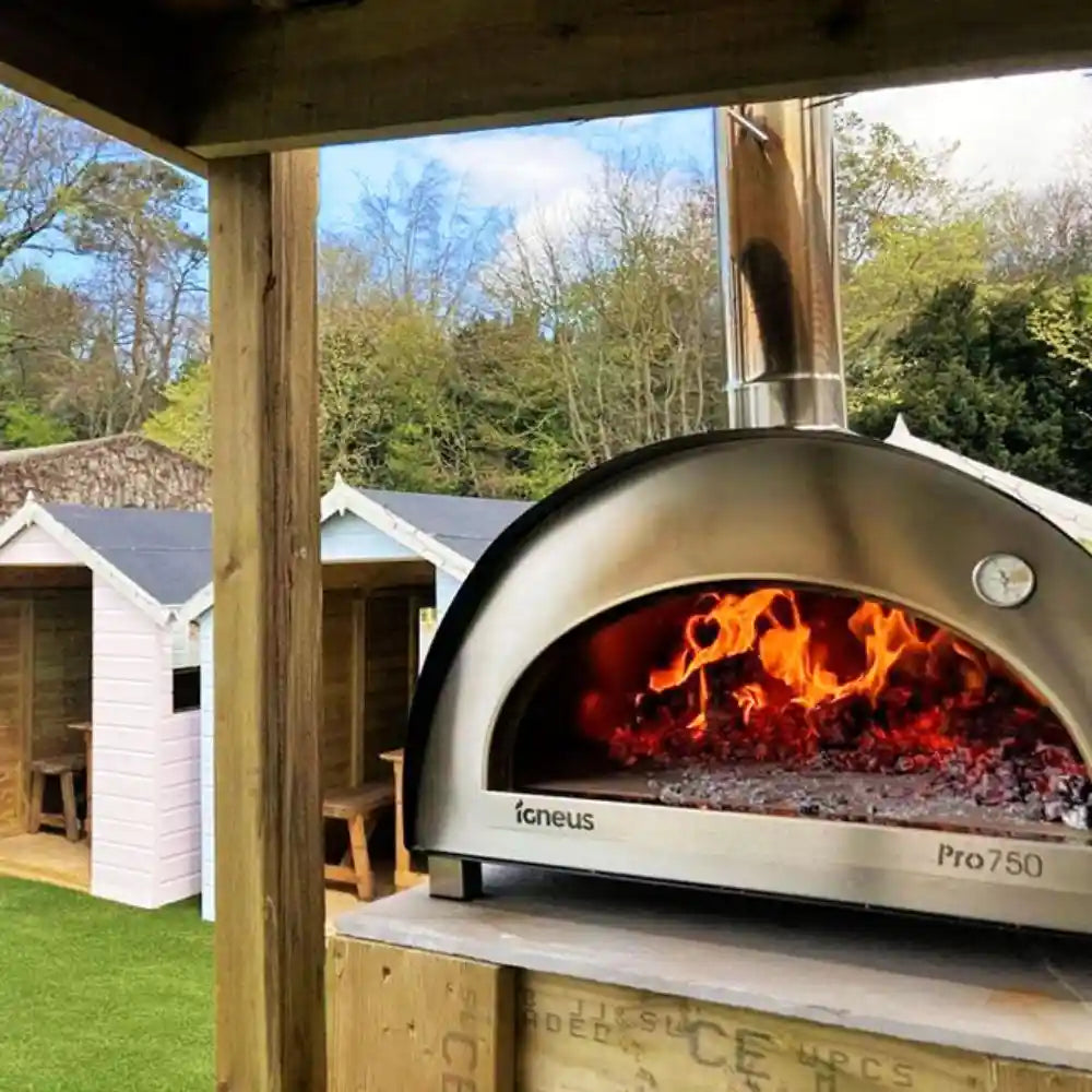 Outdoor pizza oven with flames inside, set against a wooden pergola and garden backdrop.