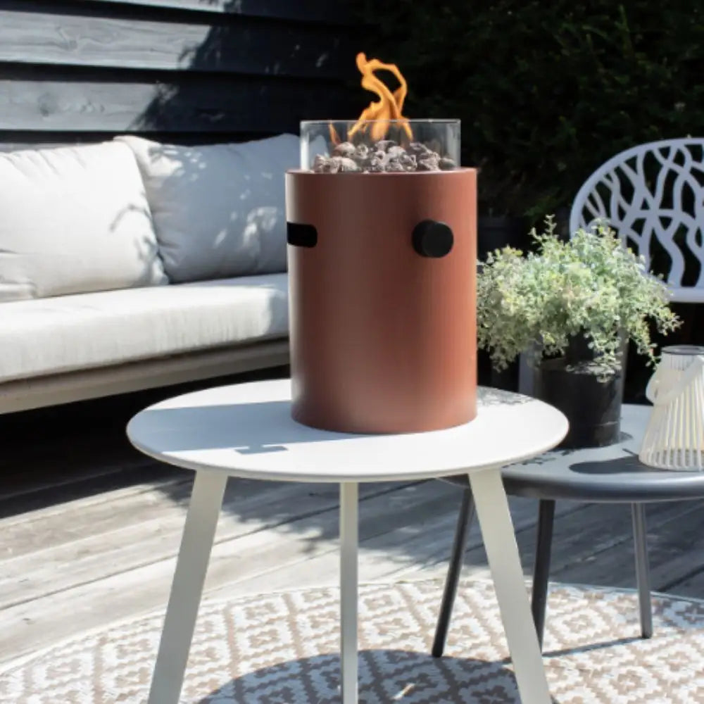 Cylindrical table top burner on a white table outdoors with a couch and plants in the background