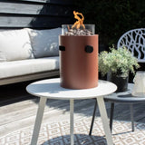 Cylindrical table top burner on a white table outdoors with a couch and plants in the background