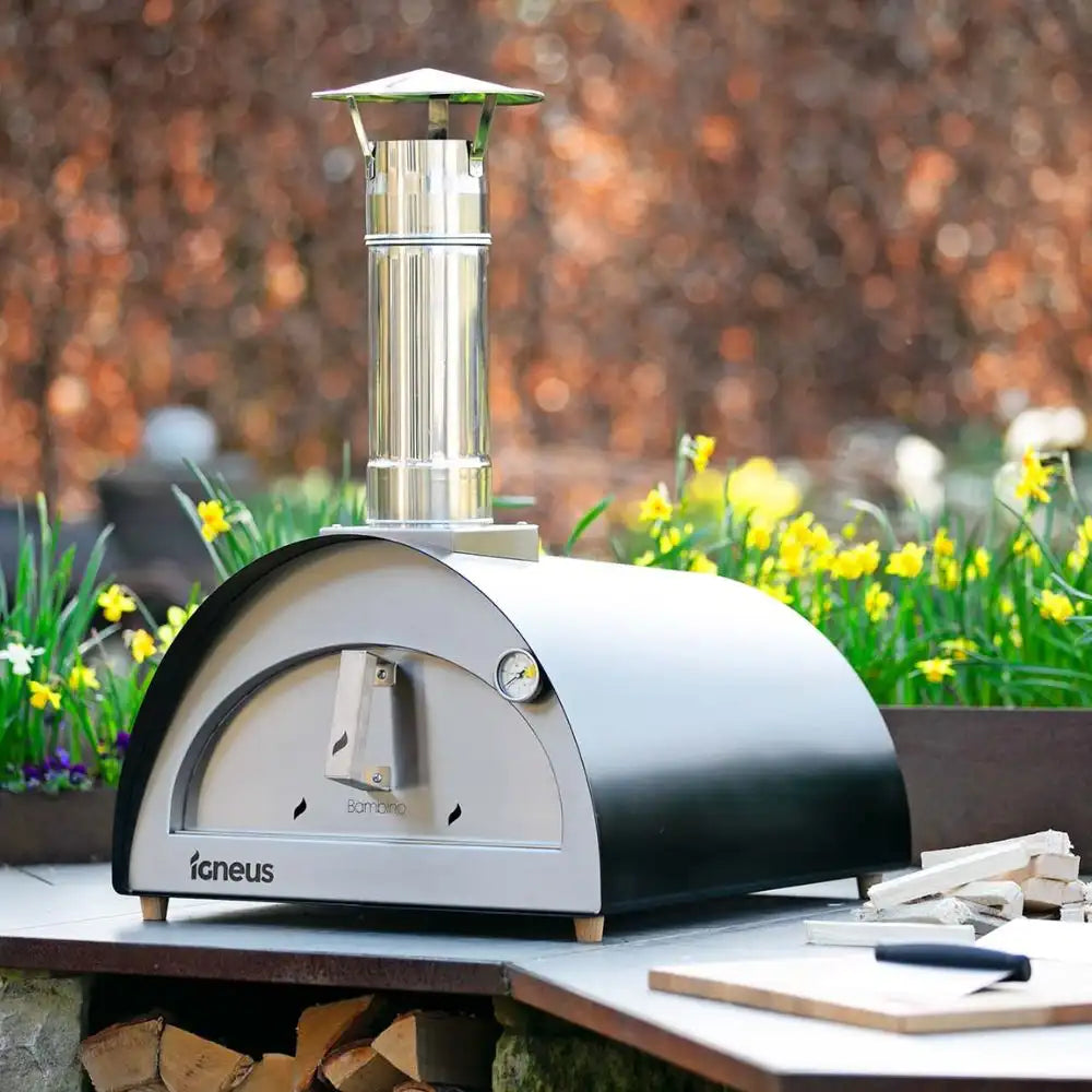 Outdoor pizza oven with 'Igneus' branding on a wooden table with flowers and logs in the background.