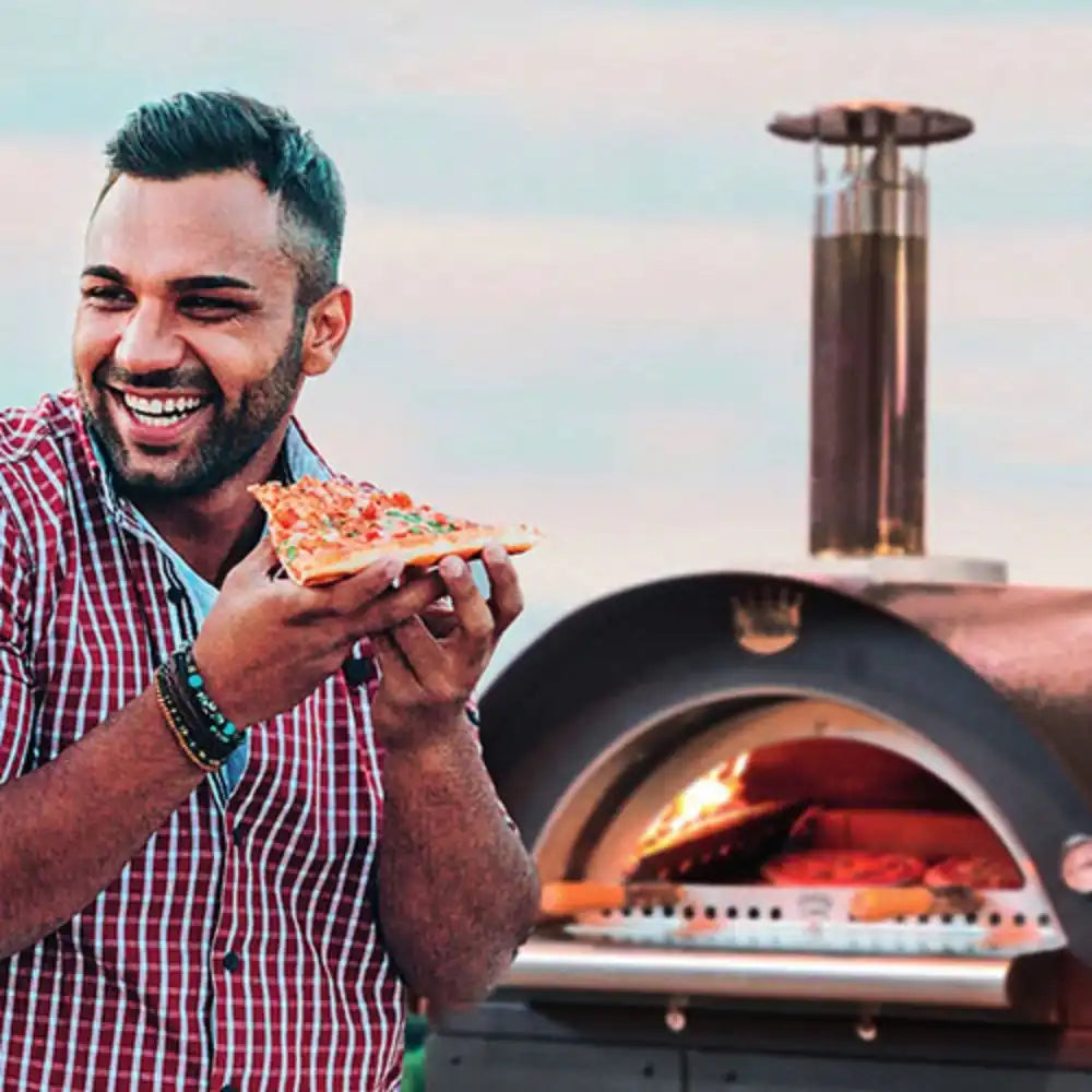 Man holding a slice of pizza in front of an outdoor pizza oven.