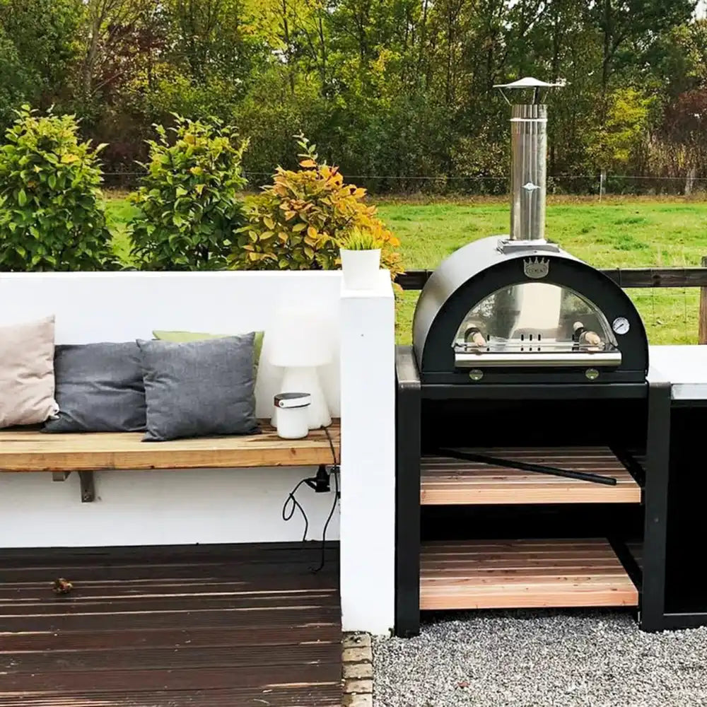 Outdoor pizza oven on a wooden deck with a bench and greenery in the background