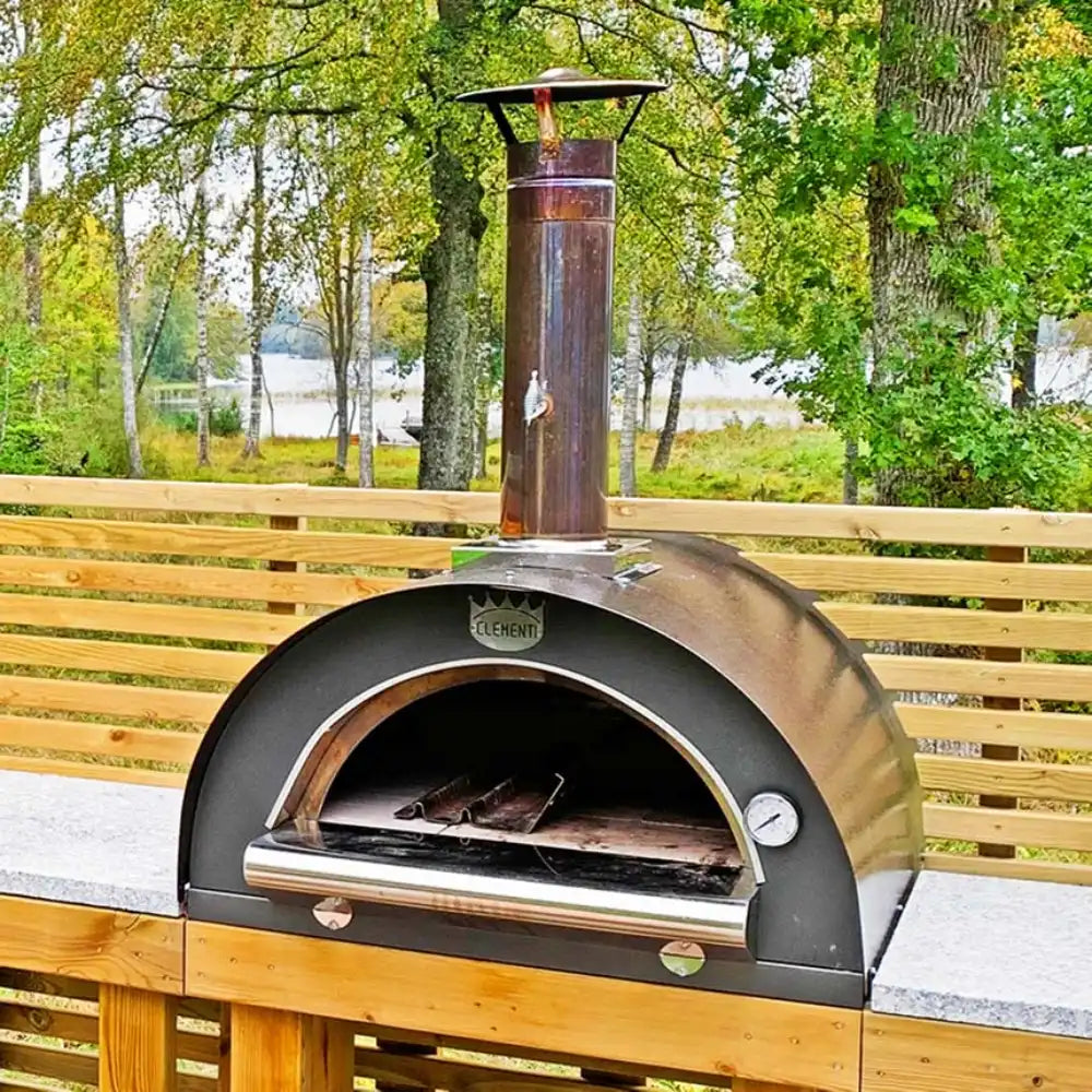 Outdoor pizza oven on a wooden deck with a scenic background of trees and water.